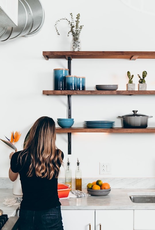 Back view of a woman in a black shirt standing at a marble kitchen counter, arranging fruit and flowers under wooden shelves with blue jars and plants.